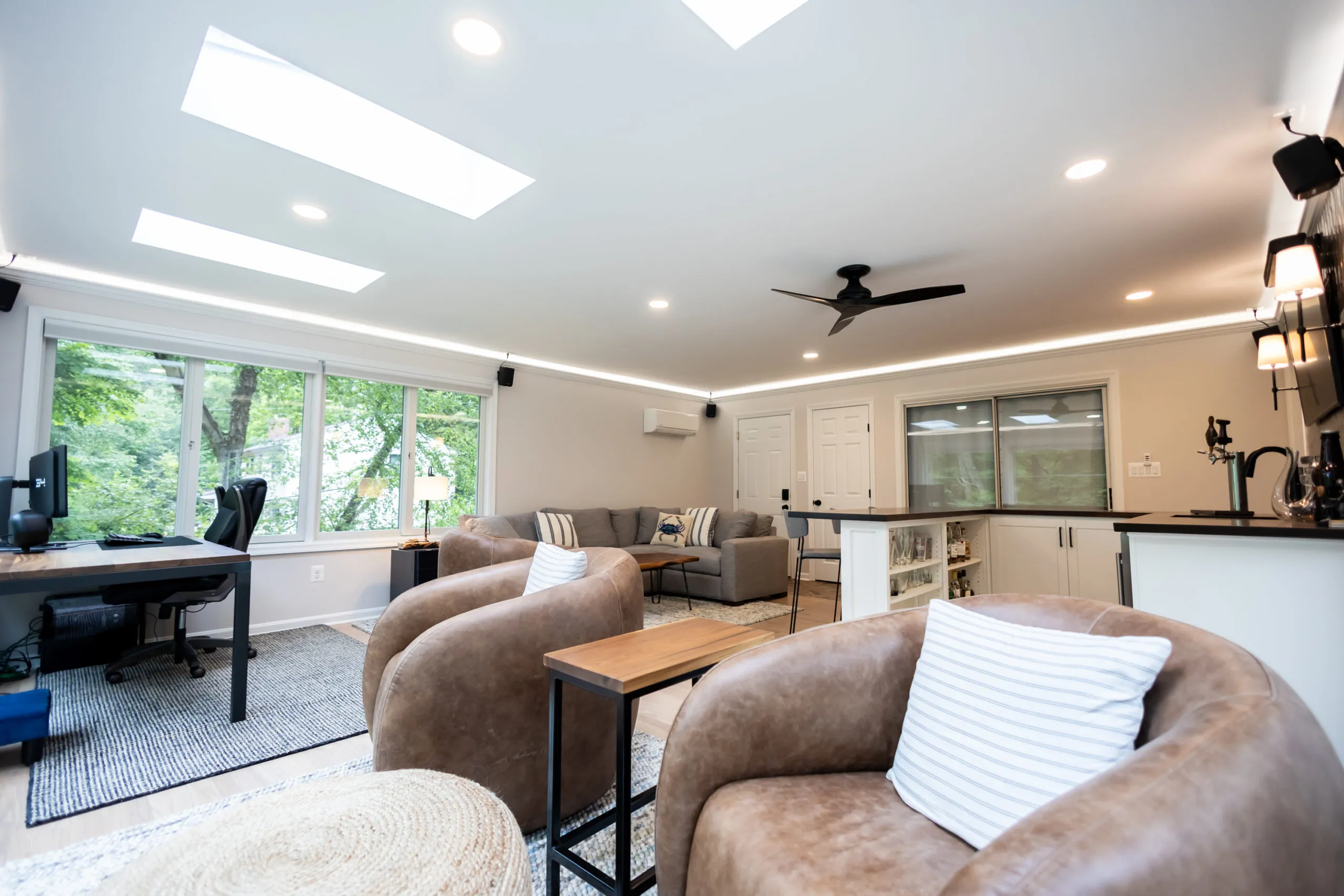 Sunlit living and office space with skylights, a wall of windows, brown leather swivel chairs, a gray sofa, and a ceiling fan