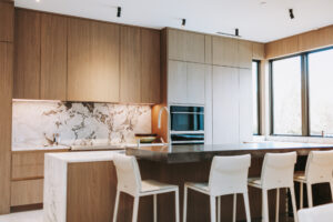 Modern kitchen with tall wooden cabinets, marble backsplash, island, and white bar stools by large windows.