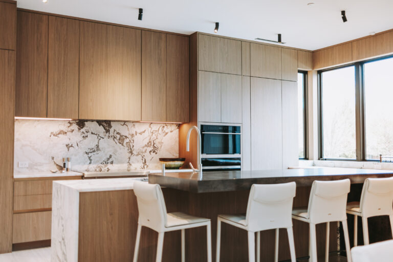 Modern kitchen with tall wooden cabinets, marble backsplash, island, and white bar stools by large windows.