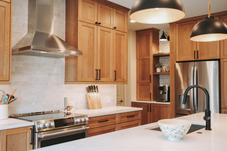 Modern kitchen with wooden cabinets, a stainless steel range hood, and a white island beneath warm pendant lights.