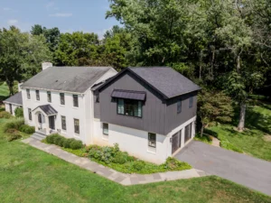 Drone view of a modern two-story house: white brick lower level, dark gray upper siding, front entry, and landscaped lawn with a stone path, trees in the background