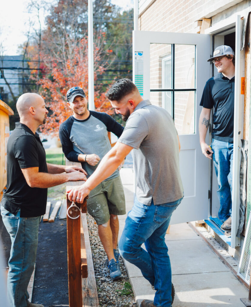Four construction workers pass a long wooden railing between them outside a house under renovation, smiling and working together.