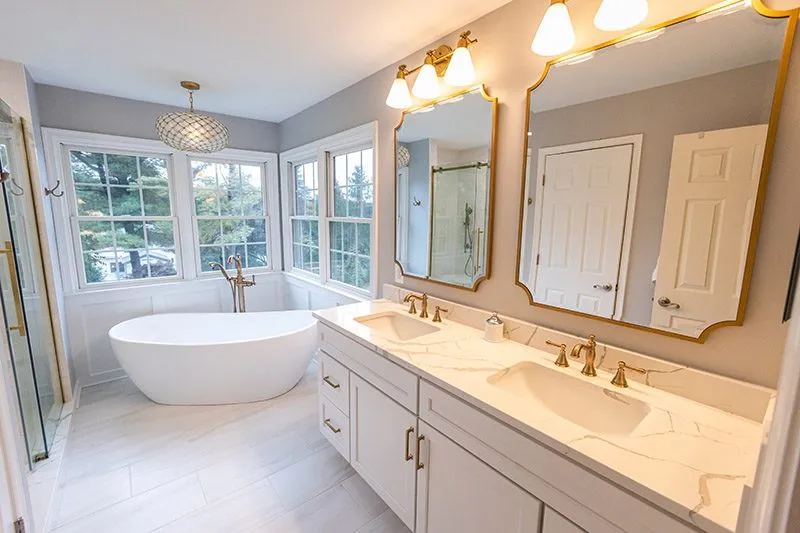Freestanding white bathtub by large bay windows in a bright bathroom with a double vanity featuring marble countertops and gold faucets, two framed mirrors, and glass shower enclosure.