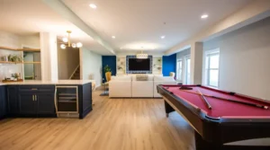 Open basement recreation space with a red pool table on the right, dark blue kitchen cabinets on the left, and a white sectional seating area facing a wall-mounted TV. The room has light wood floors and blue accent walls.