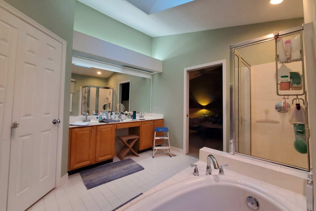 Bathroom with a double-sink vanity, green walls, and large mirror reflecting a shower stall and open doorway to a dark room beyond.