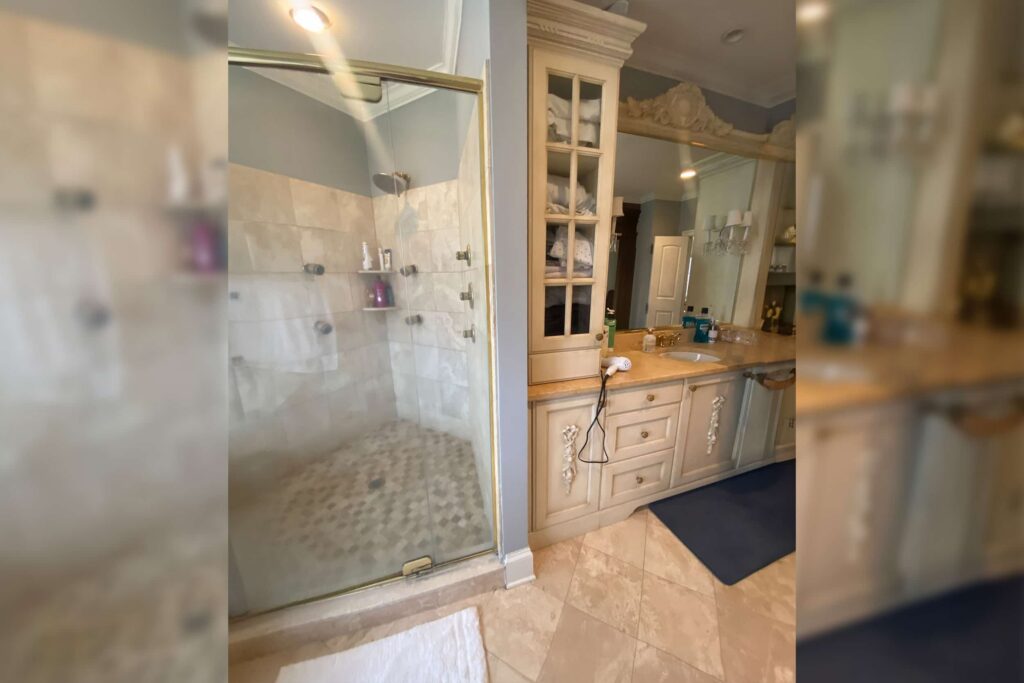 Bathroom with glass-enclosed shower on left, beige tile, and a wooden vanity with a marble countertop on the right; open cabinet with towels above the counter.