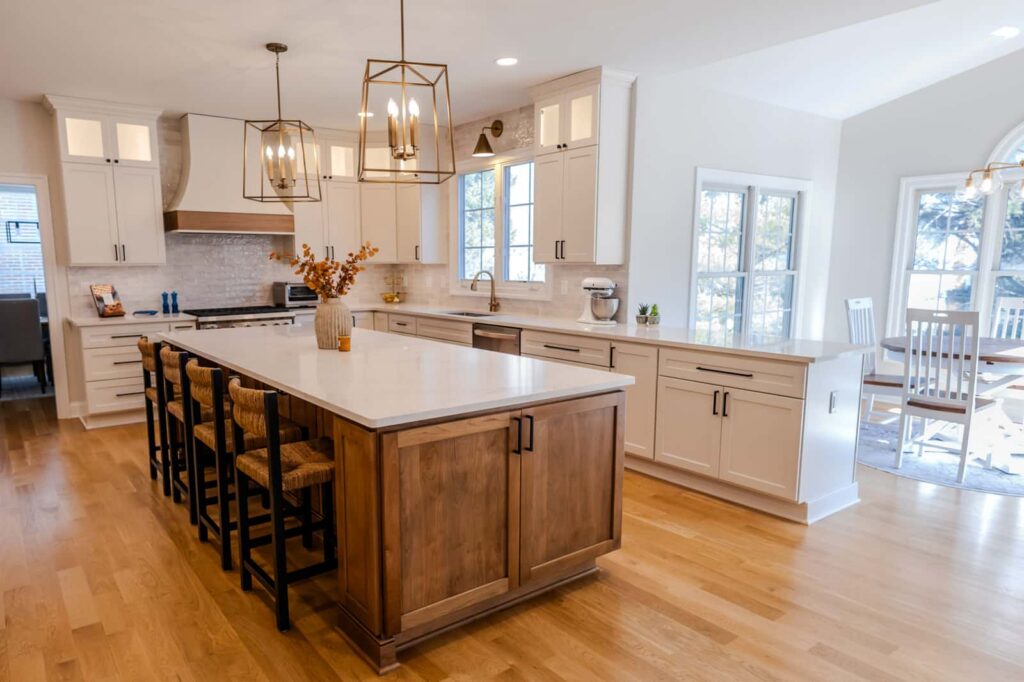 Bright open kitchen with white cabinets, a large white island, wood base and six woven stools, and geometric gold pendant lights.