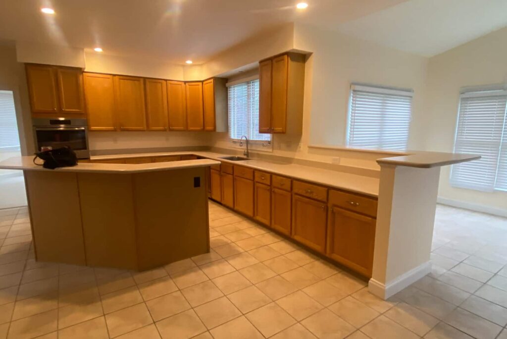 U-shaped kitchen with light wood cabinets, white countertops, a curved island, and a built-in oven; tile floor and window blinds.
