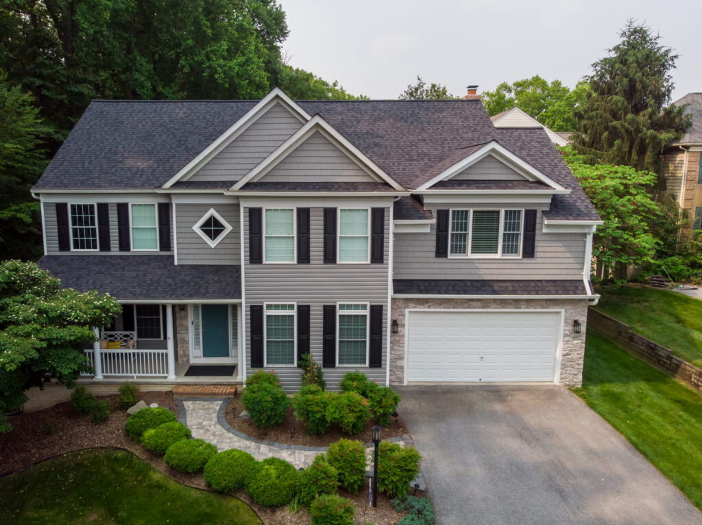 A two-story gray suburban house with black shutters, a two-car garage, and a manicured front yard (drone view).