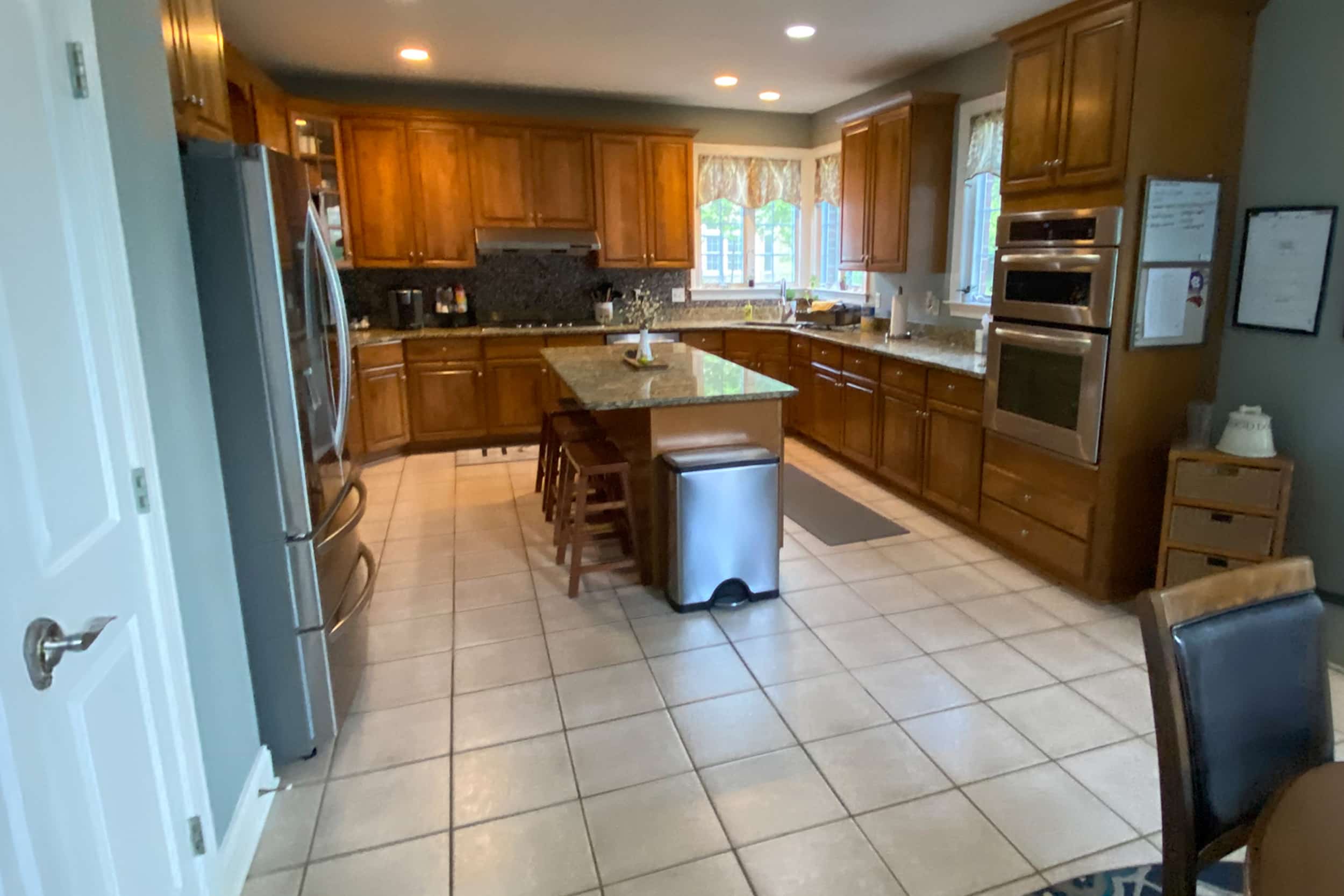 L-shaped kitchen with wooden cabinets, granite countertops, and an island at center. Stainless steel fridge on the left and a wall oven on the right.