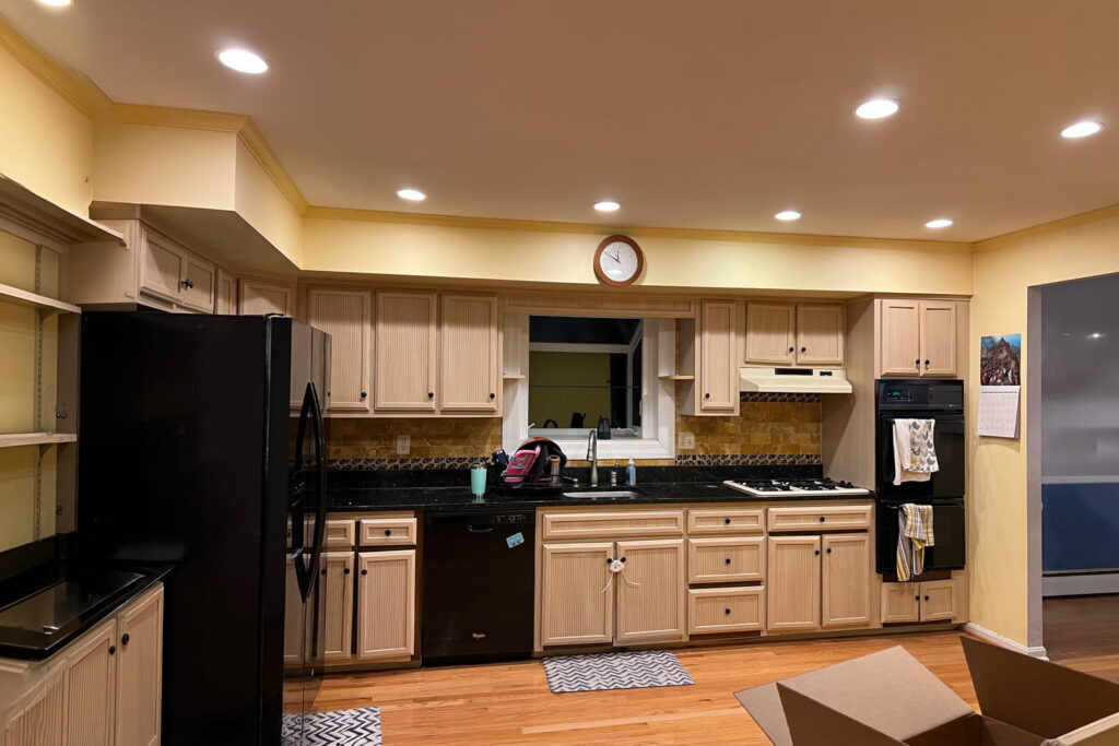 U-shaped kitchen with light wood cabinets, black countertops, and stainless steel appliances; a window above the sink and a wall clock above the cabinets.