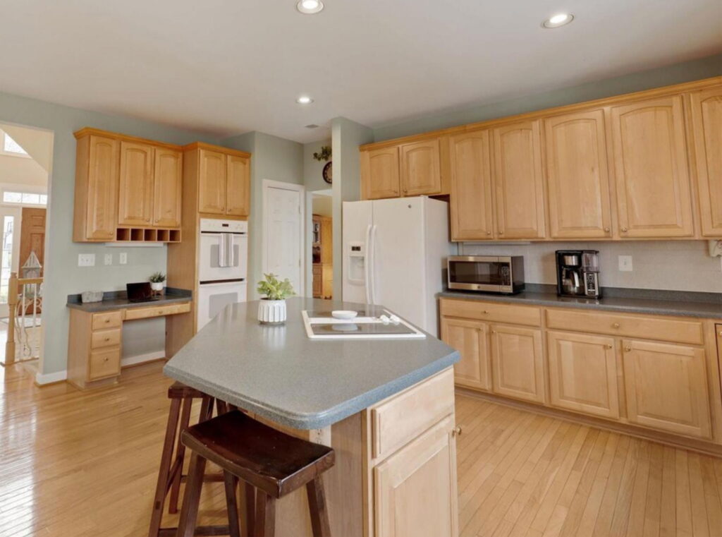 Bright kitchen with light wood cabinets, grey countertops, and a center island with two dark-stained stools.