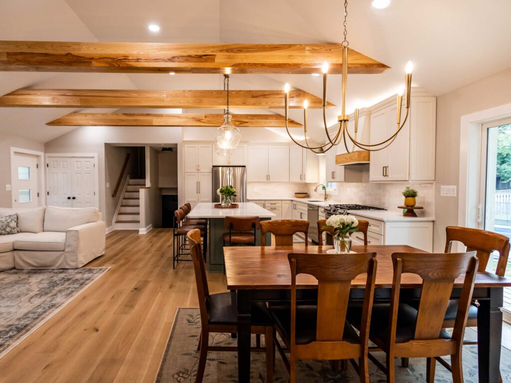 Open-concept kitchen with white cabinets, stainless appliances, wooden ceiling beams, and a gold chandelier over the island.