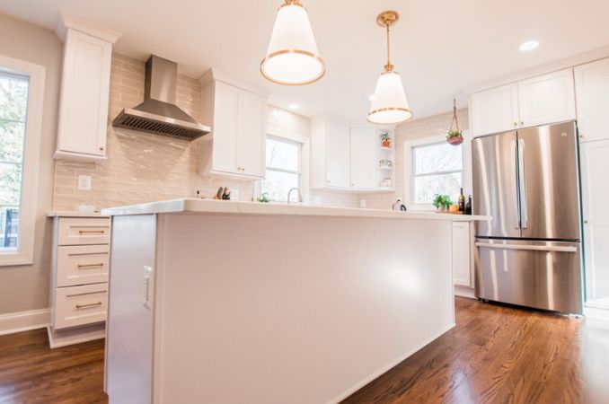 Bright modern kitchen with a large white island, pendant lights, white cabinets, and a stainless steel refrigerator.