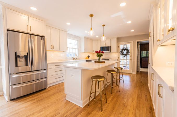 Bright white kitchen with an island, three gold-bar stools, and a stainless steel French-door refrigerator on oak flooring.
