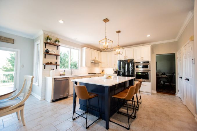 Bright white kitchen with a large central island, three brown bar stools, and pendant lights over the island. A stainless steel fridge and double wall ovens are visible.