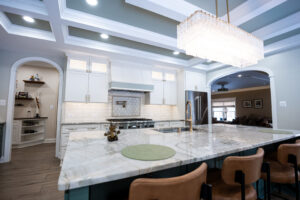 Bright kitchen with marble island, white cabinets, and a large crystal chandelier overhead while arches open to the living area.