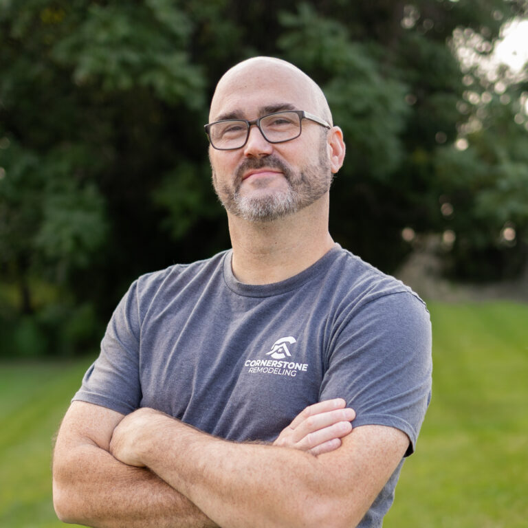 Man with glasses and a short beard stands outdoors with arms crossed, wearing a gray Cornerstone Remodeling shirt.