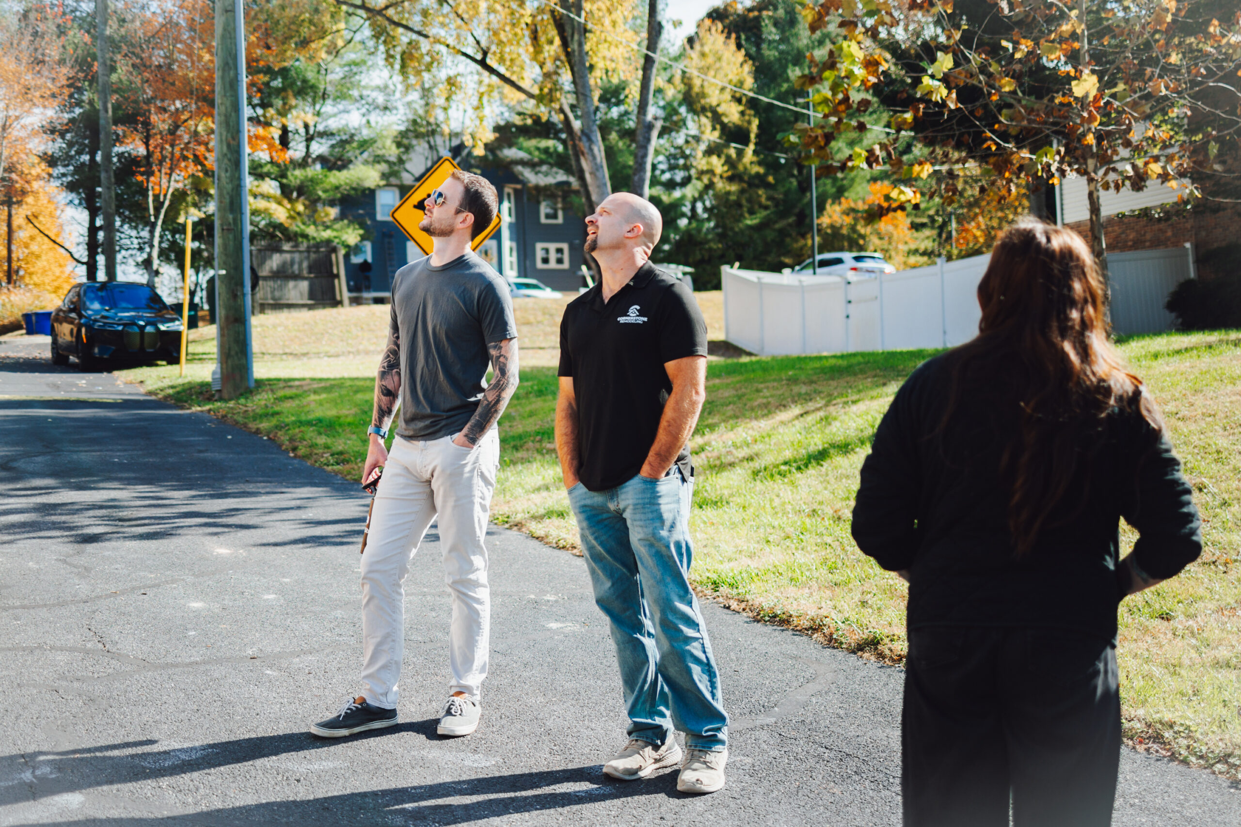 Three people stand on a suburban street looking upward, autumn trees in the background and a person with back to camera in foreground.