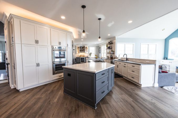Open-concept kitchen with white cabinets, a dark gray island, stainless steel double ovens, pendant lights over the island, wood-look flooring, and large windows bringing in natural light.