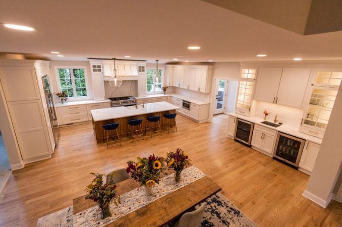 Open-concept white kitchen with a large island and four navy stools, stainless appliances, and a wooden dining table with flower arrangements in the foreground.